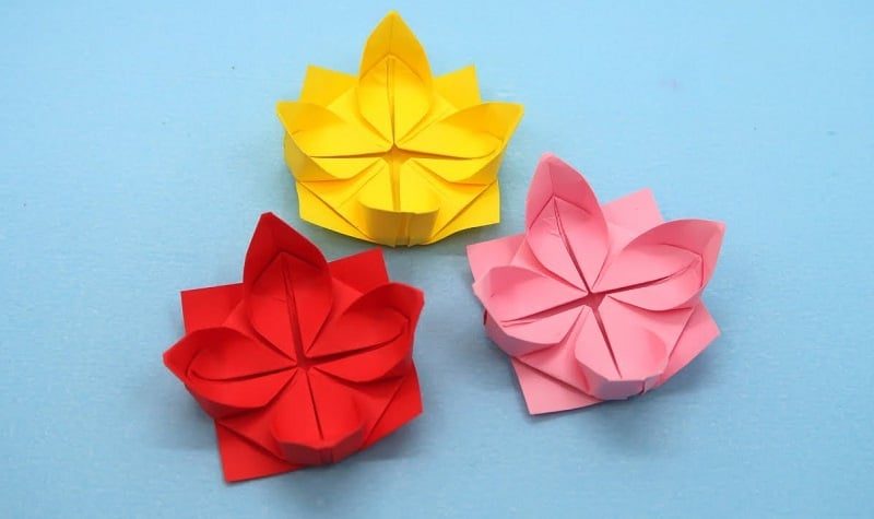 Trois fleurs de lotus en papier origami (jaune, rouge, rose) posées sur fond bleu