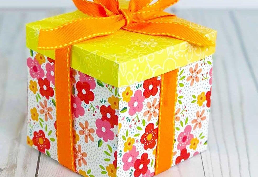 Boîte explosive en papier fleuri fermée avec un nœud en ruban orange, posée sur une table en bois