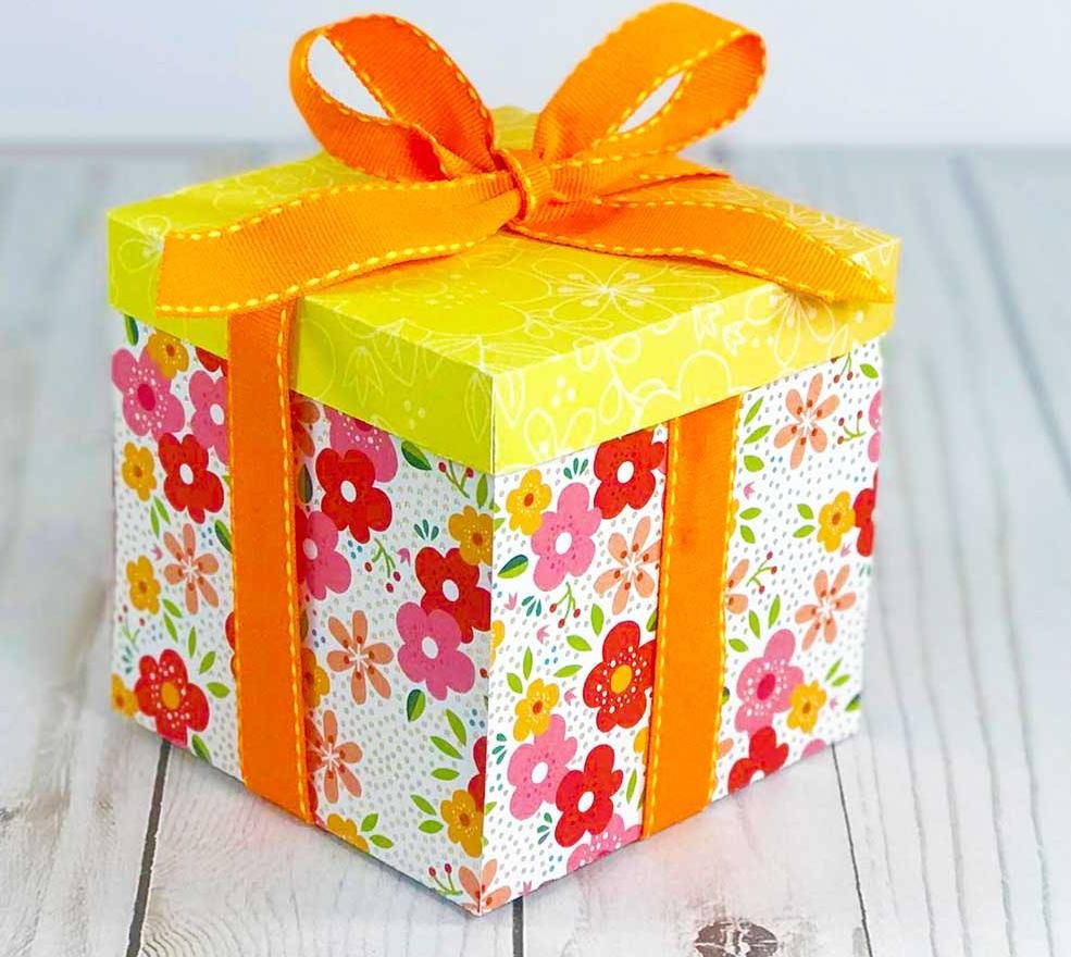 Boîte explosive en papier fleuri fermée avec un nœud en ruban orange, posée sur une table en bois