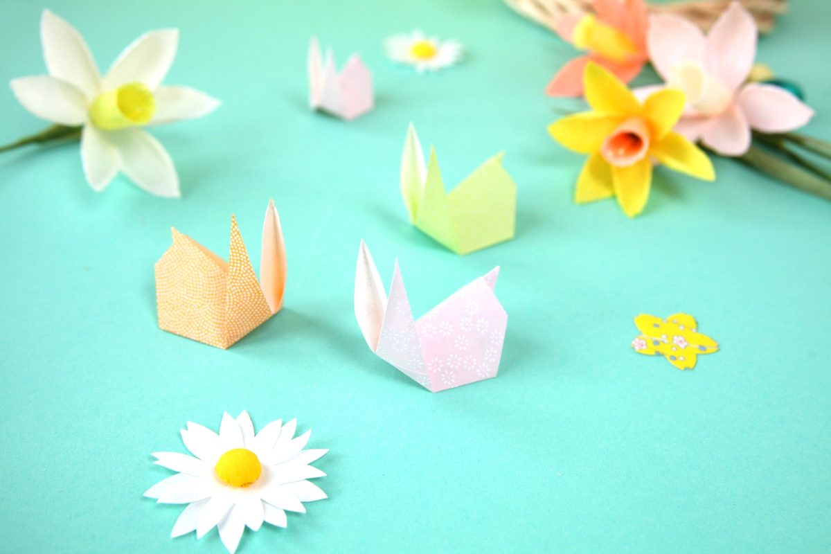 Petits lapins en origami pliés dans du papier japonais pastel, posés parmi des fleurs en papier
