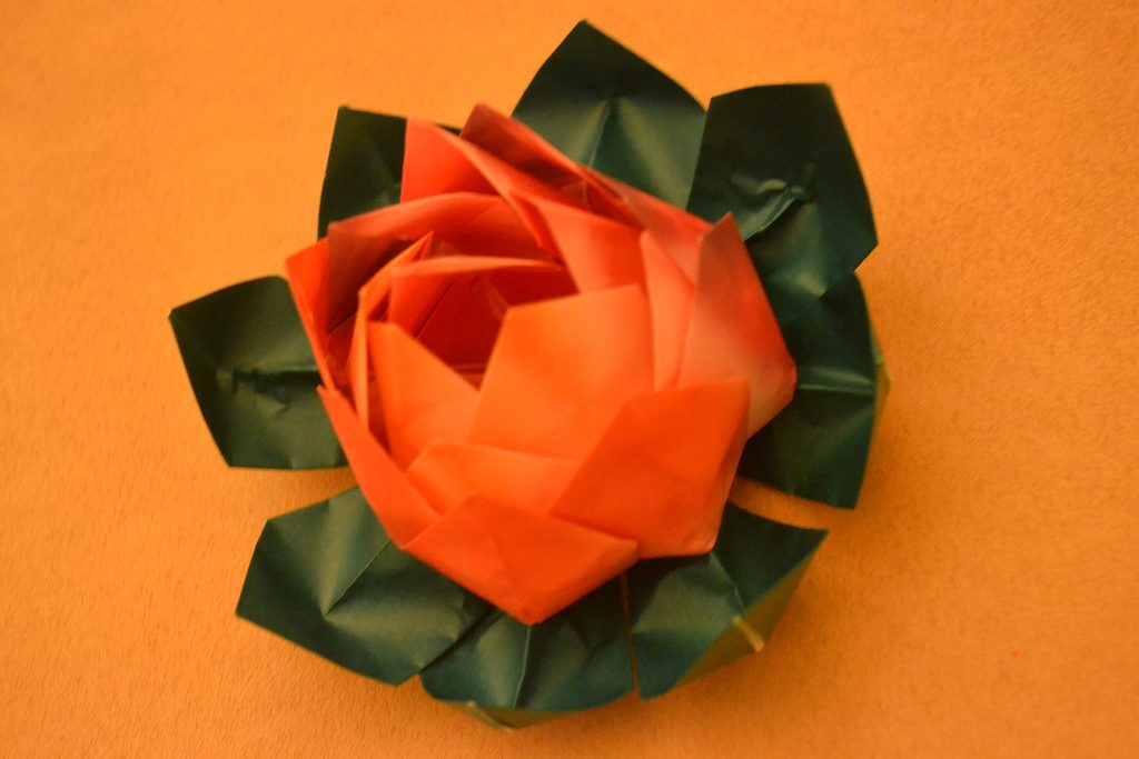 Fleur de lotus en origami rouge posée sur des pétales verts, sur fond orange
